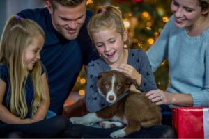 A happy family with two young girls and a puppy in front of a Christmas tree, sharing a joyful moment.